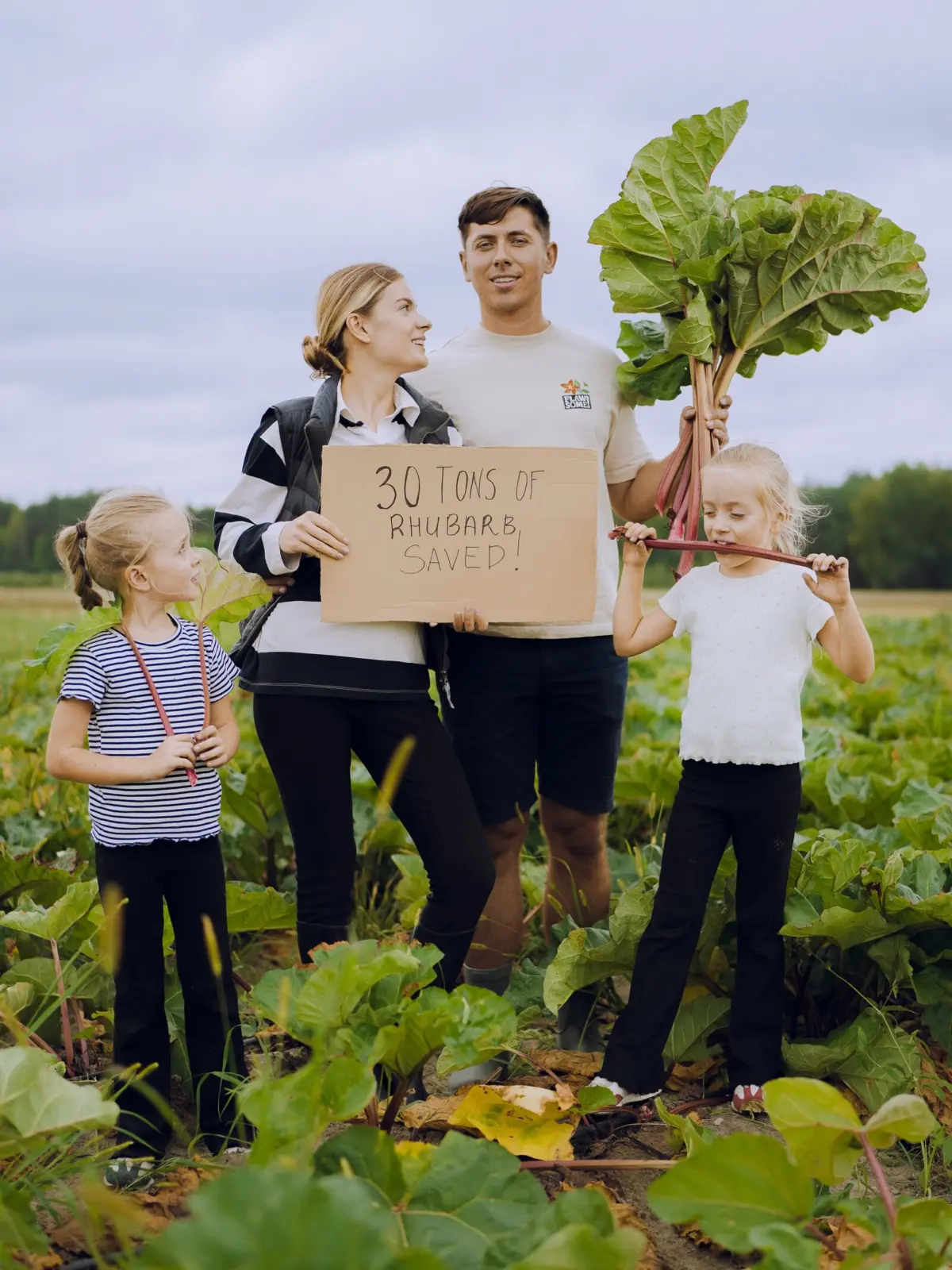 Karina and Maciek of Flawsome Drinks standing in a rhubarb field with their two daughters, holding a handwritten cardboard sign reading 30 tons of rhubarb saved